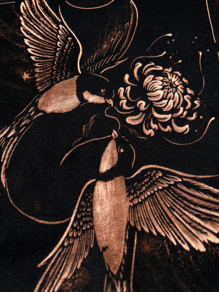 Close-up of a dark fabric or print featuring two birds and a large flower in warm metallic beige tones against a black background. Hand-painted bleach art wall hanging of The Lovers tarot card, featuring two birds carrying a flower, wood dowel frame with black fringe