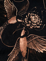 Close-up of a dark fabric or print featuring two birds and a large flower in warm metallic beige tones against a black background. Hand-painted bleach art wall hanging of The Lovers tarot card, featuring two birds carrying a flower, wood dowel frame with black fringe