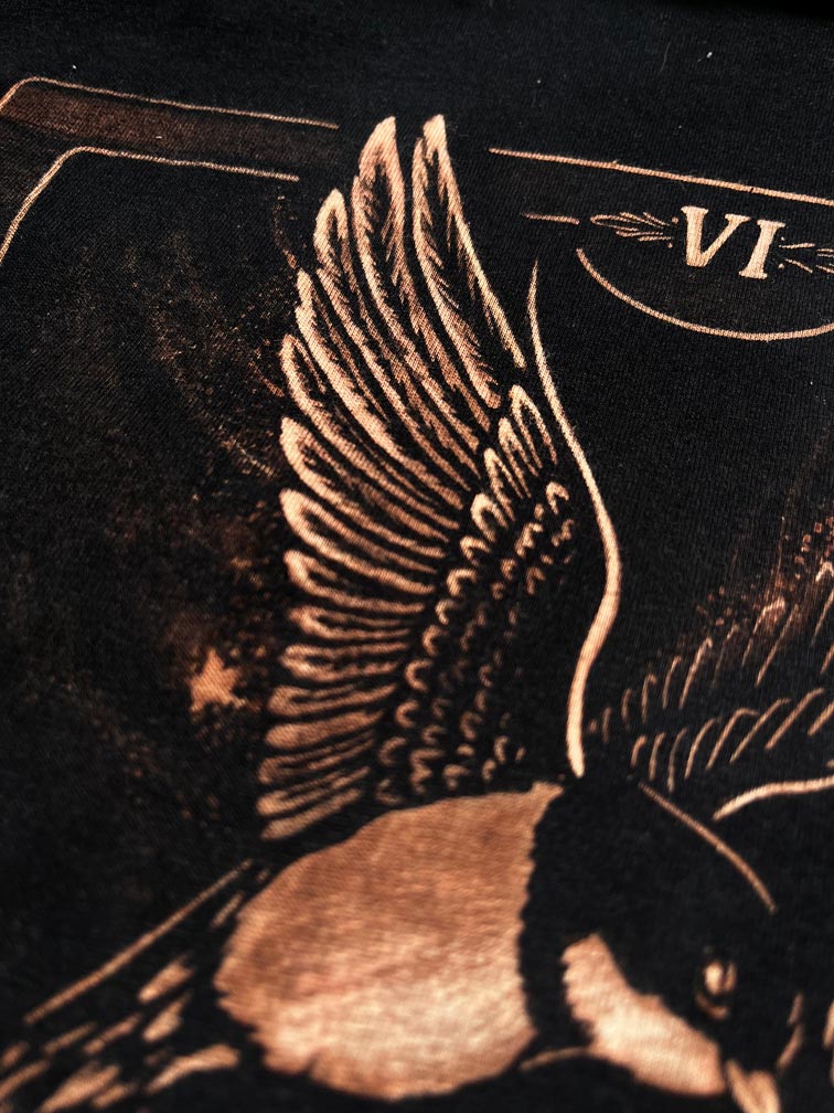 Close-up of a black garment featuring a detailed copper-toned graphic of a bird with spread wings and the Roman numeral VI near the top.