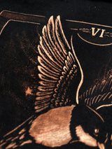 Close-up of a black garment featuring a detailed copper-toned graphic of a bird with spread wings and the Roman numeral VI near the top.