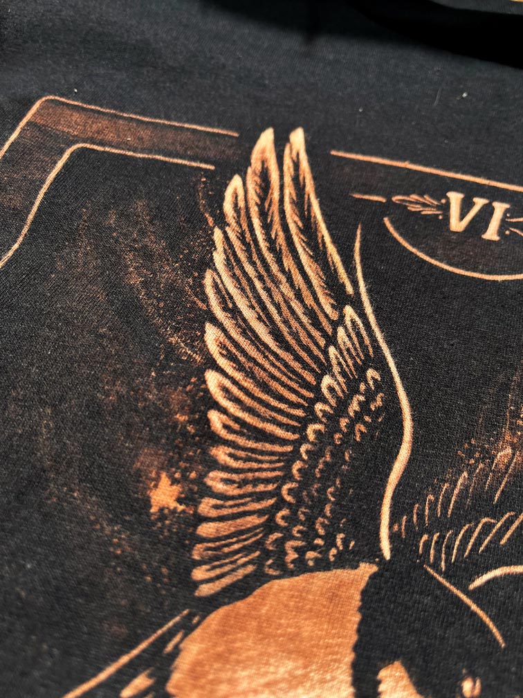 Close-up of a black graphic print featuring a large winged figure in metallic copper or bronze tones, with part of a card-like design and the Roman numeral VI visible. Hand-painted bleach art wall hanging of The Lovers tarot card, featuring two birds carrying a flower, wood dowel frame with black fringe