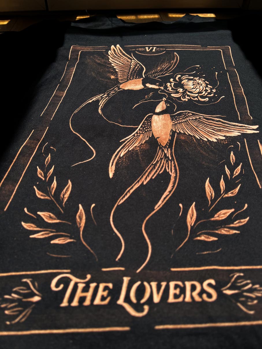 A black t-shirt featuring a tarot card-style graphic of two birds holding a flower, with the text 'THE LOVERS' and Roman numeral 'VII' at the top, shown laid flat on a dark surface.