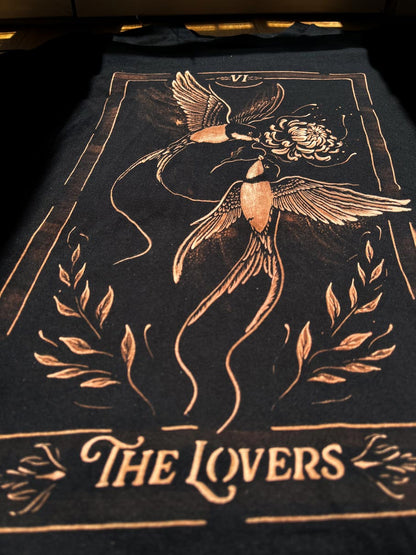 A black t-shirt featuring a tarot card-style graphic of two birds holding a flower, with the text 'THE LOVERS' and Roman numeral 'VII' at the top, shown laid flat on a dark surface.