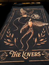 A black t-shirt featuring a tarot card-style graphic of two birds holding a flower, with the text 'THE LOVERS' and Roman numeral 'VII' at the top, shown laid flat on a dark surface.