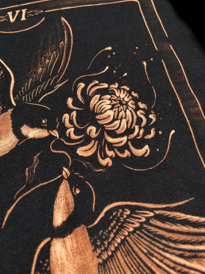 Close-up of a black bleach artfeaturing two birds and a large flower in a warm metallic copper or bronze tone. Tarot card design t-shirt VI: the lovers