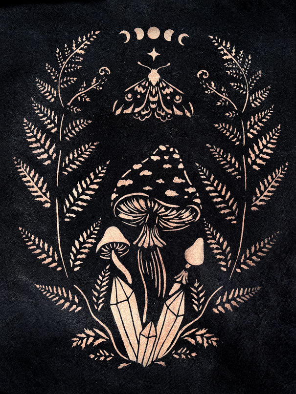 A stencil design painted with bleach on fabric featuring a stylized illustration of mushrooms, fern leaves, a crescent moon and a star motif at the top