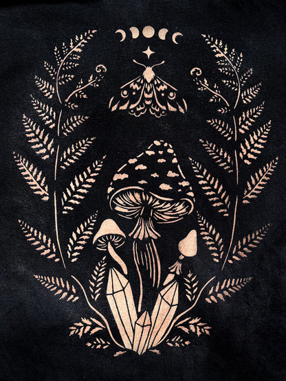 A stencil design painted with bleach on fabric featuring a stylized illustration of mushrooms, fern leaves, a crescent moon and a star motif at the top