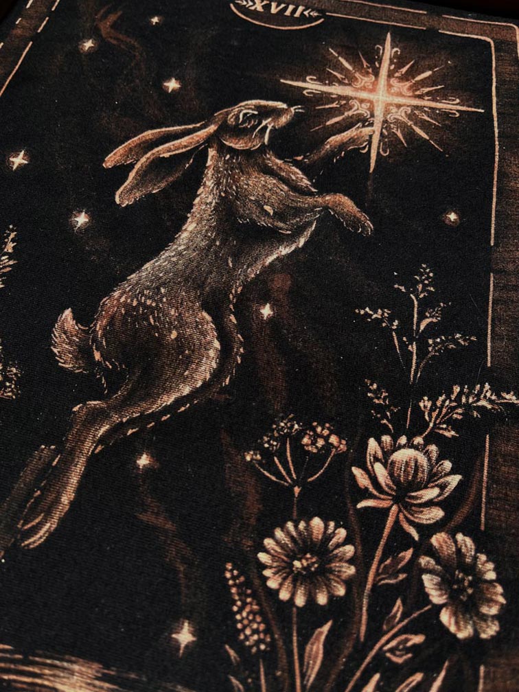 A close-up of a dark tarot card illustration featuring a hopping rabbit reaching toward a glowing star, with floral details and the tarot card design VII the star