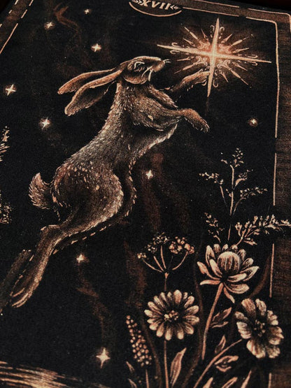 A close-up of a dark tarot card illustration featuring a hopping rabbit reaching toward a glowing star, with floral details and the tarot card design VII the star