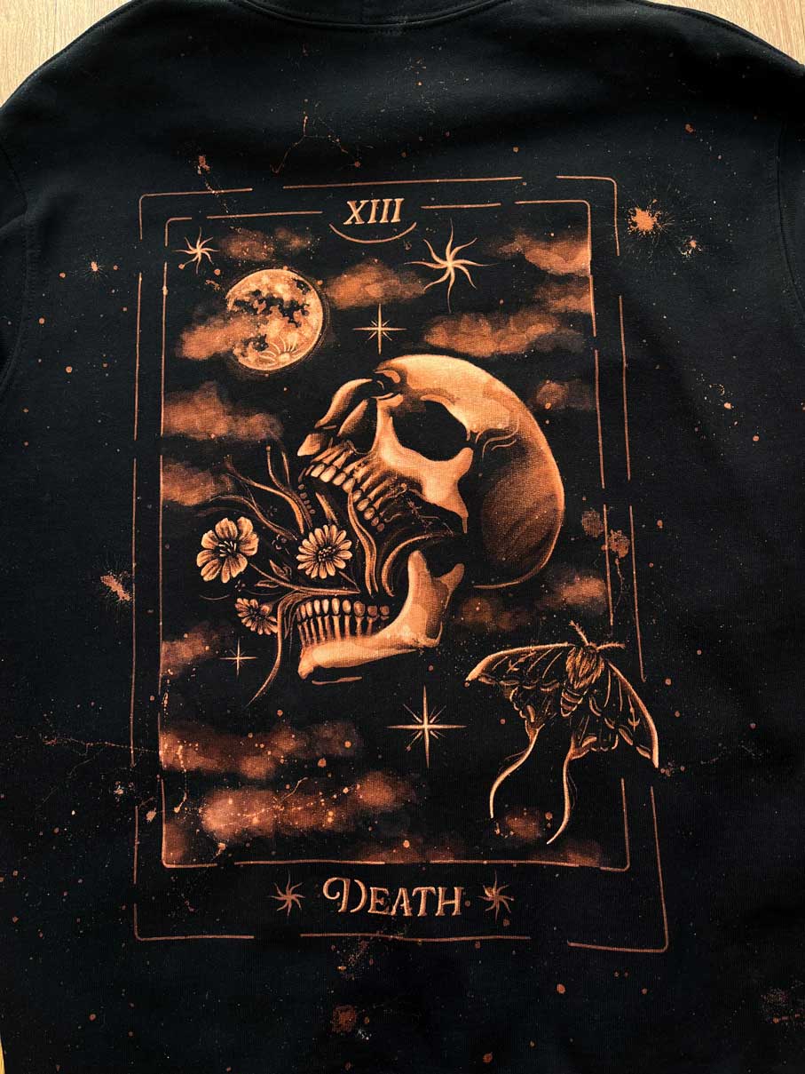 A close-up of a black garment featuring a large tarot card-style graphic on the front. The design shows a skull, moon, stars, flowers, a moth, and the word 'DEATH' in a brown and tan palette.