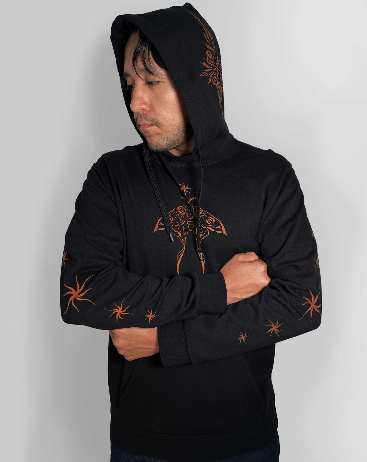 A man wearing a black pullover hoodie with orange bleach art on the sleeves and chest, posed against a light gray background.