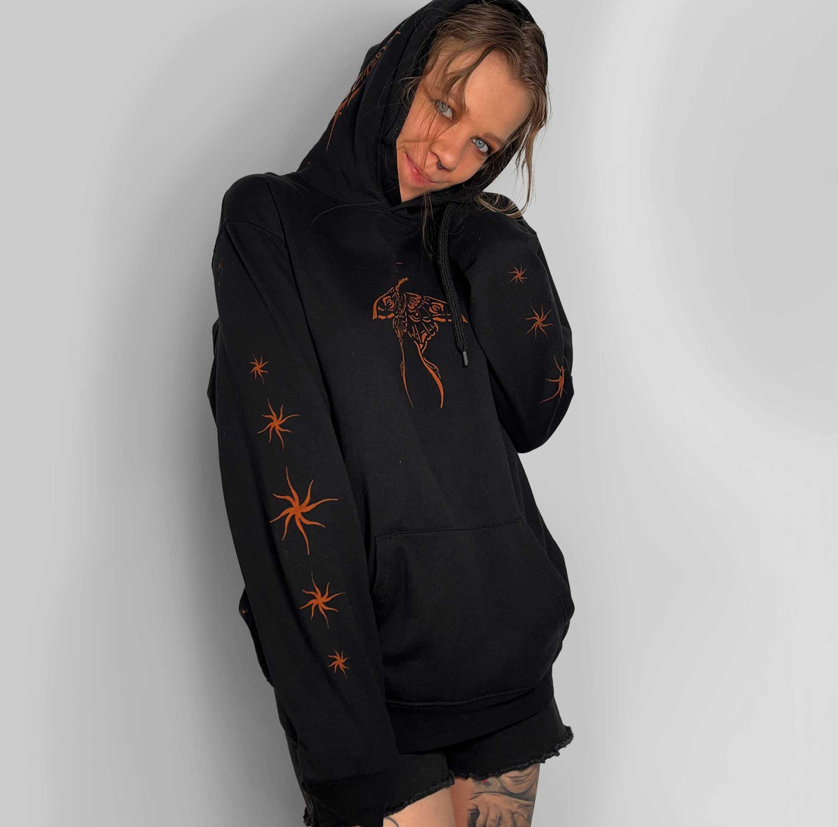 A person wearing a black pullover hoodie with orange bleach art, including a moth on the chest and starburst motifs on the sleeves, posed against a light gray wall.