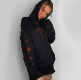 A person wearing a black pullover hoodie with orange bleach art, including a moth on the chest and starburst motifs on the sleeves, posed against a light gray wall.
