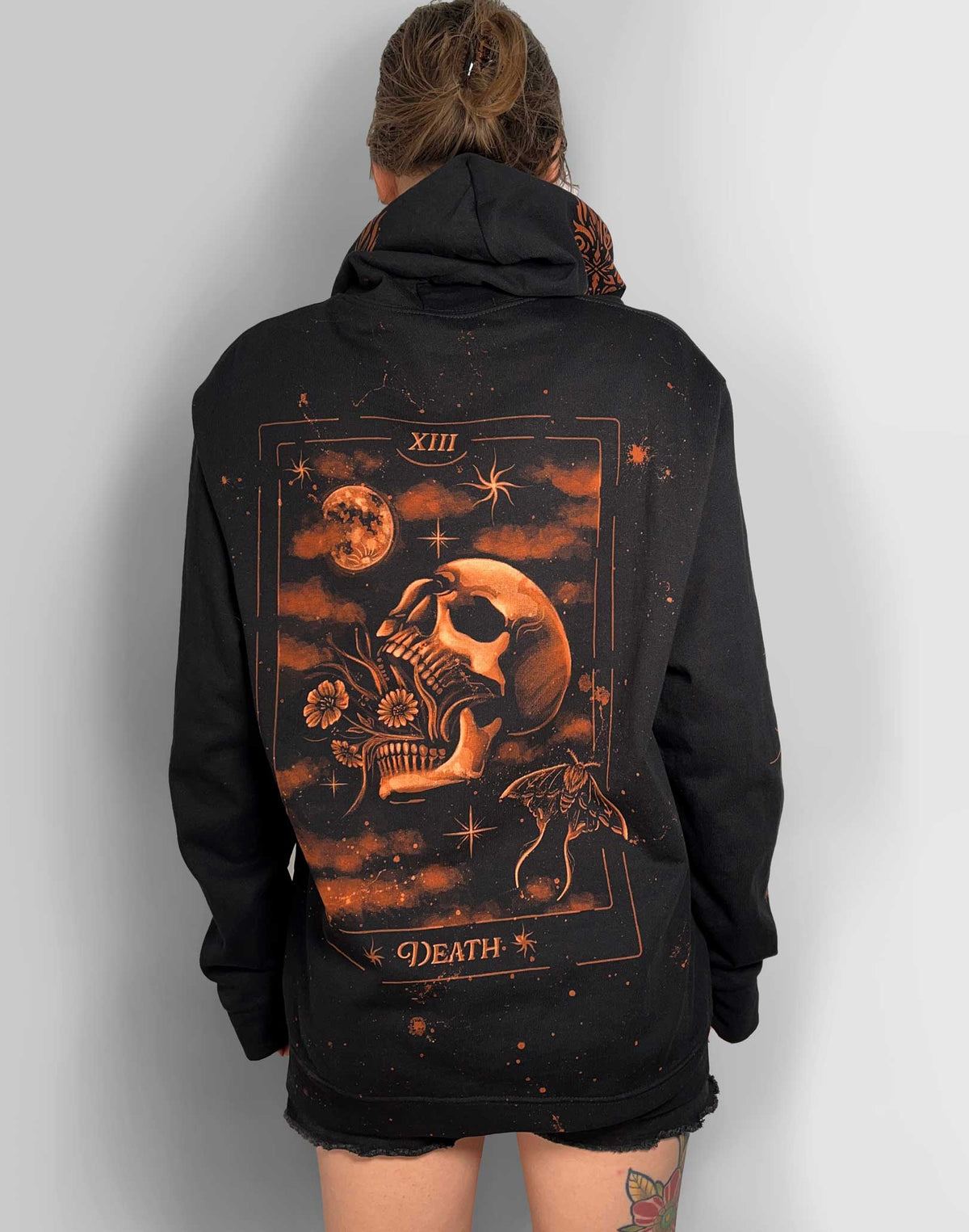 A person viewed from the back wearing an oversized black hoodie with a large tarot-style Death graphic in orange and brown tones across the back, against a plain light gray wall.