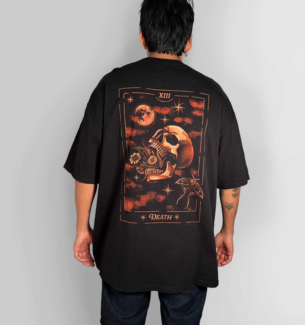A person shown from the back wearing an oversized black T-shirt with a large tarot-style graphic on the back featuring a skull, moon, stars, and the word 'DEATH' in orange and brown tones, against a light gray studio background.