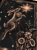 A close-up of a dark tarot card illustration featuring a hopping rabbit reaching toward a glowing star, with floral details and the Roman numeral XVIII at the top.