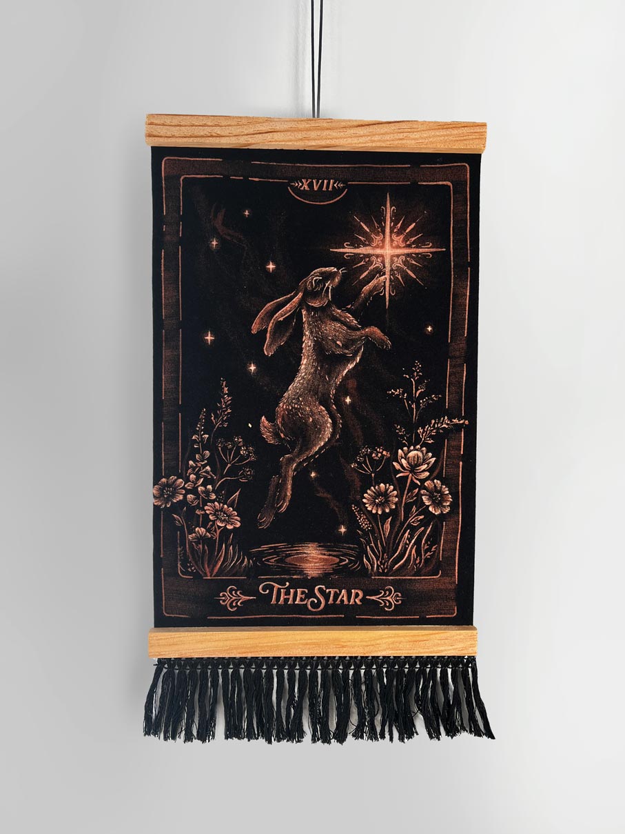 A vertical black tapestry or wall hanging with a wooden top and bottom bar, featuring a brown rabbit under a glowing star and the text 'XVII' and 'THE STAR'. A black fringe hangs from the bottom. Hand-painted tarot card inspired wall art 
