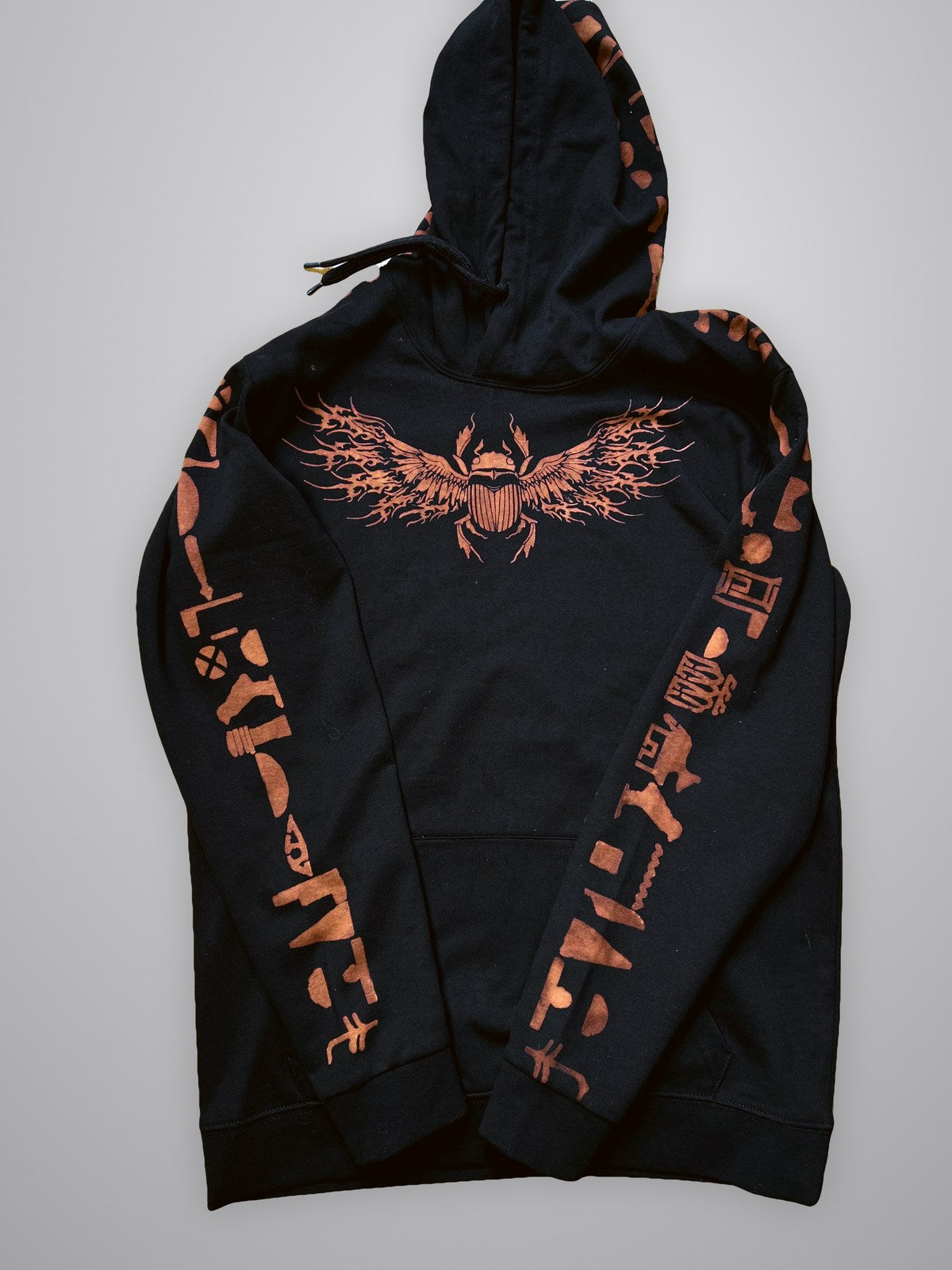 Flat lay of the Anubis hoodie front with a winged scarab on the chest and hieroglyphs down both sleeves and the hood
