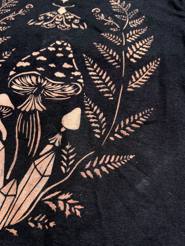 Close-up of a black fabric surface painted with bleach botanical and mushroom pattern, including fern leaves, stems, and mushroom illustrations.