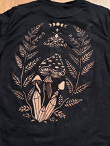 A close-up of a black t-shirt showing the back graphic, which features bleach painted mushrooms, crystals, ferns, a moth, and moon phases.