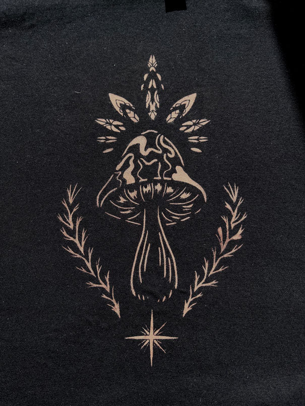 A close-up of a black fabric surface featuring a bleach stencil art graphic of a mushroom centered within leafy and laurel-like branches, with a small starburst below.