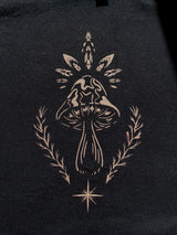 A close-up of a black fabric surface featuring a bleach stencil art graphic of a mushroom centered within leafy and laurel-like branches, with a small starburst below.