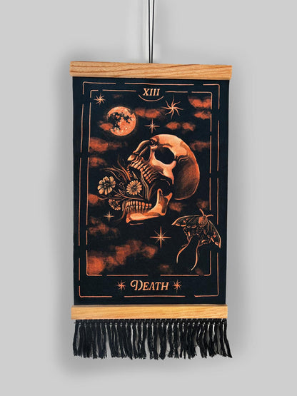 A vertical black tapestry banner featuring a tarot-style 'Death' illustration with a skull, moon, stars, and butterfly, suspended from a light wood rod with black fringe at the bottom against a light gray background.