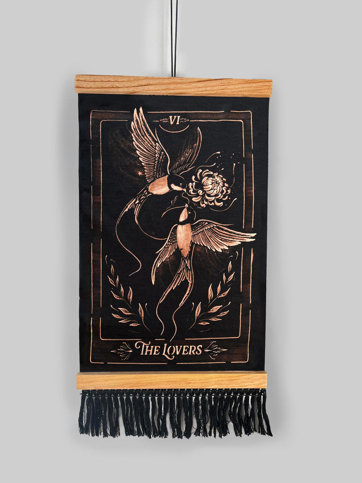 A vertical black wall tapestry featuring a bronze-toned engraved design of 'The Lovers' tarot card, suspended from a light wood dowel with black fringe at the bottom against a solid gray background. Hand-painted bleach art wall hanging 