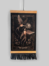 A vertical black wall tapestry featuring a bronze-toned engraved design of 'The Lovers' tarot card, suspended from a light wood dowel with black fringe at the bottom against a solid gray background. Hand-painted bleach art wall hanging 