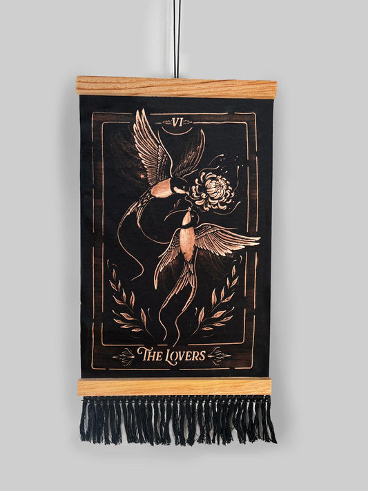 A vertical black wall tapestry featuring a bronze-toned engraved design of 'The Lovers' tarot card, suspended from a light wood dowel with black fringe at the bottom against a solid gray background. Hand-painted bleach art wall hanging 